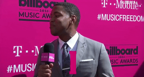 A man in a grey suit speaks into a T-Mobile microphone on the pink carpet at the 2016 Billboard Music Awards, smiling as pink confetti falls, attracting media attention. #MUSICFREEDOM and Billboard Music Awards signs are visible in the background.