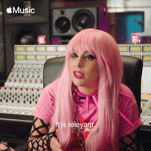 A person with long pink hair and bold make-up sits in a music studio, wearing a pink top and fishnet sleeves, speaking with the caption It is relevant. The Apple Music logo is in the top left corner, hinting at social media secrets for shareable content.