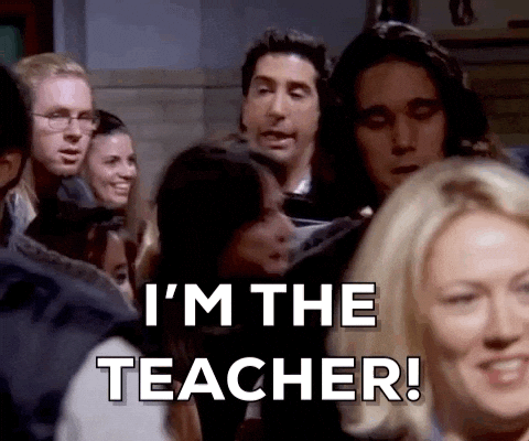A man in a crowded room shouts I’M THE TEACHER! using storytelling as people gather round, some smiling and laughing in the background.