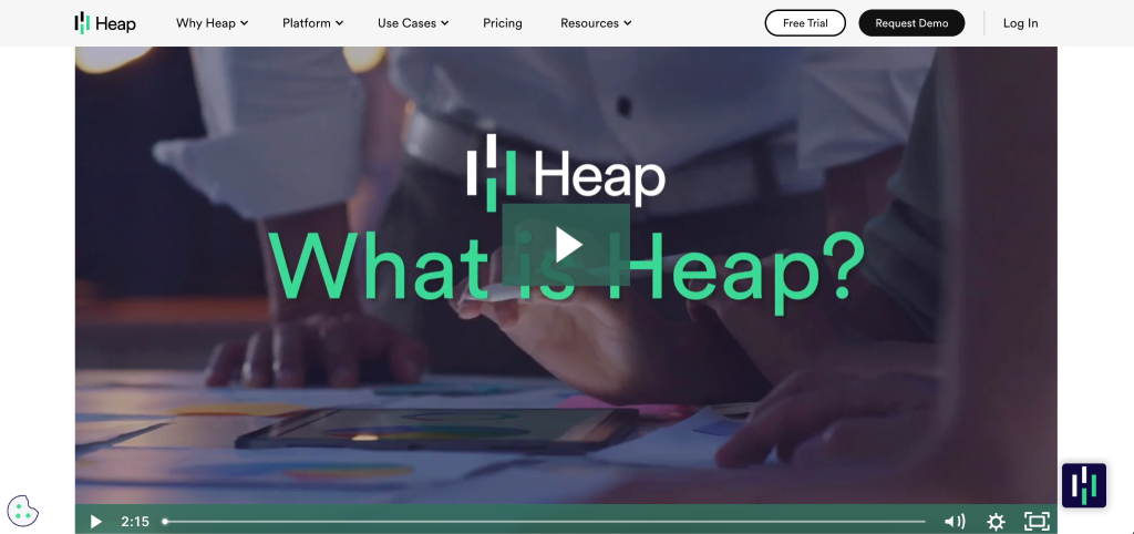A paused video on the Heap website shows people collaborating over documents on a table. Overlaid text reads “What is Heap?” with Heap’s logo, navigation menus, and sleek website design visible at the top of the screen.